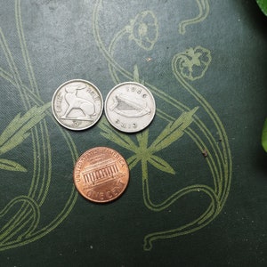 May include: Three coins on a green surface with a floral pattern. The coins are a silver Irish penny with a hare, a silver Irish penny with a harp, and a copper US penny with the Lincoln Memorial.