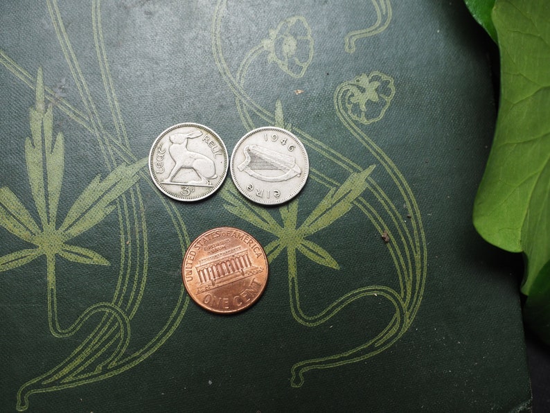 May include: Three coins on a green surface with a floral pattern. The coins are a silver Irish penny with a hare, a silver Irish penny with a harp, and a copper US penny with the Lincoln Memorial.
