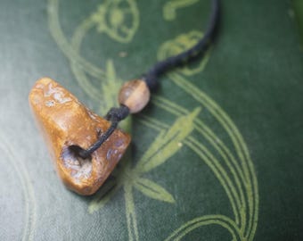 May include: A handcrafted pendant necklace featuring a unique, light brown stone with a hole. The stone is attached to a black cord with a wooden bead. The necklace rests on a green surface with a floral design.