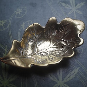 May include: A metallic, gold-toned dish shaped like an oak leaf. The surface of the dish features a detailed, embossed vein pattern that mimics the natural structure of a leaf, with a polished finish that reflects light across its curved edges.