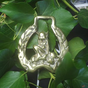 May include: A polished brass horse brass featuring a central embossed figure of a seated imp or goblin-like creature surrounded by a decorative border with a textured, leaf-like pattern and a rectangular loop at the top for attachment.