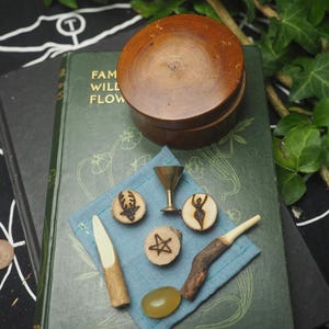 May include: A wooden box with a lid sits atop a green book with the words "FAM WILL FLOW". A blue square cloth holds small wooden charms, a brass goblet, and a wooden spoon. The charms feature various symbols.