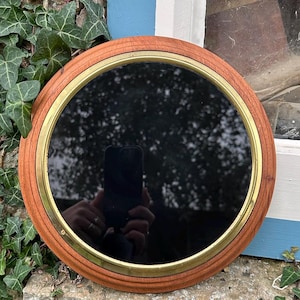 May include: Round wooden mirror with a brass-colored metal band. The mirror has a dark reflective surface and is set against a stone wall with green ivy. The frame is made of light brown wood.