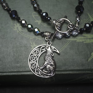 May include: A silver-toned necklace featuring a howling wolf pendant set within a crescent moon design. The necklace has two strands of dark beads with a toggle clasp. The pendant is detailed with Celtic knotwork and a five-pointed star.