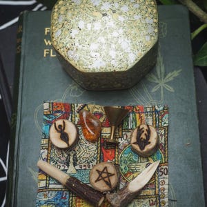 Puede incluir: Una colección de objetos místicos, que incluye una caja hexagonal decorativa con un diseño floral, amuletos de madera, un cristal y un pequeño cáliz de latón. Los objetos están dispuestos sobre una tela estampada colorida y un libro antiguo.