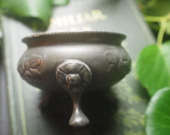 May include: A small, round, dark metallic bowl with a matte finish, featuring three ornate, curved legs and embossed leaf patterns along the exterior. The rim has a beaded texture, and the overall design is consistent with a vintage metal vessel.