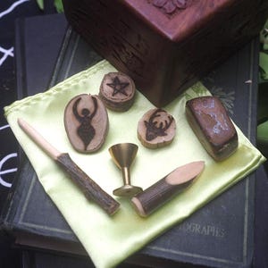 Puede incluir: Una colección de pequeños objetos rituales que incluye tres discos de madera con símbolos quemados de una diosa, una estrella y un ciervo, un pequeño cáliz de latón, una varita de madera, una estaca de madera y una piedra marrón lisa.