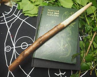 May include: A handcrafted wooden wand with a light-colored tip rests on a vintage book titled "Familiar Wild Flowers." The wand is brown with a natural wood grain and decorative knots. Green leaves and a black and white patterned cloth surround the book.