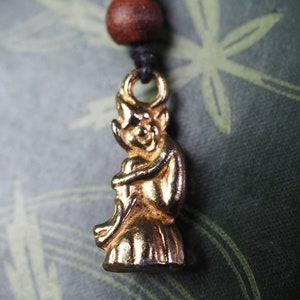 May include: A gold-coloured pendant featuring a detailed figurine of a woman in a flowing gown. The pendant is attached to a black cord with a brown wooden bead.