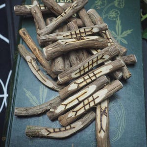 May include: A collection of wooden rune sticks, each etched with intricate symbols, rests on a vintage green book. The natural wood tones contrast with the dark etched designs, creating a rustic and mystical aesthetic. The runes are likely used for divination or spiritual practices.