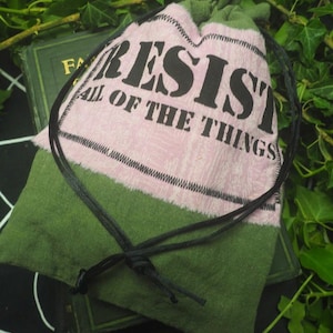 May include: A drawstring pouch made of olive green fabric with a rectangular pink patch sewn onto the front. The patch features the black text RESIST ALL OF THE THINGS. A thin black cord is tied around the top of the pouch to secure the opening.
