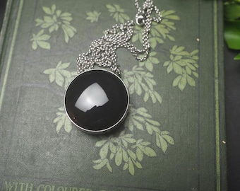 May include: A round, polished black cabochon pendant set in a simple silver-toned metal frame, suspended from a matching silver-toned ball chain necklace. The smooth, dark stone features a prominent white light reflection on its curved surface.
