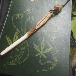 Puede incluir: Un colgante de varita mágica de madera hecha a mano con una punta clara y un mango marrón oscuro. La varita está suspendida de un cordón negro y descansa sobre un libro verde con diseños florales y de hojas. Hojas verdes en el fondo.