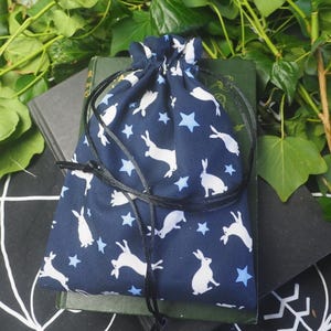 May include: A navy blue drawstring bag featuring a white rabbit and blue star pattern. The bag is made of fabric and has a black drawstring closure. The bag is resting on a green book.