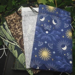 May include: Four rectangular fabric drawstring pouches featuring distinct patterns: a dark blue bag with gold suns, moons, and butterflies; a light grey bag with white rabbits and floral motifs; a brown bag with a dark geometric print; and a green bag with leaves.