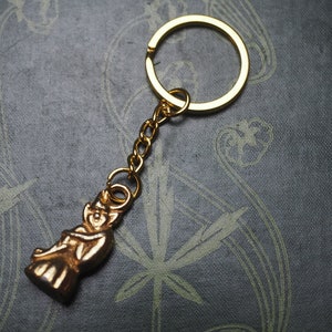 Puede incluir: Un llavero dorado con una pequeña figura detallada de una persona. El llavero está unido a un anillo de metal dorado.