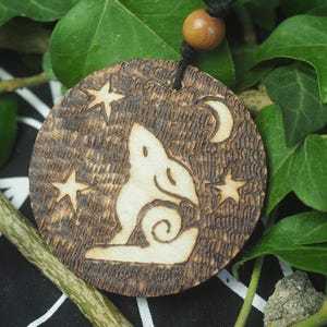 May include: A round wooden pendant with a carved design of a howling wolf, stars, and a crescent moon. The pendant is attached to a black cord with a wooden bead. The pendant is set against a background of green leaves.