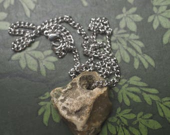 May include: A silver-toned metal chain necklace featuring a natural, irregularly shaped tan and grey stone pendant. The stone has a smooth, circular hole drilled through the top section, allowing the chain to pass directly through the rock.