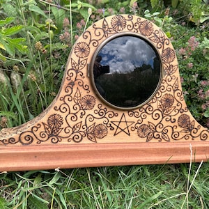 Peut inclure: Une horloge décorative en bois avec un centre rond en verre foncé. L'horloge présente des motifs floraux et de feuilles complexes, ainsi qu'une étoile à cinq branches. L'horloge est posée sur une base en bois et possède un cadre de couleur laiton autour du verre.