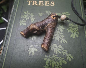 May include: A Y-shaped dark brown wooden pendant with a textured bark surface and a small hole at the top. A black cord passes through the hole, secured by a single light-colored round bead. The pendant rests on a green book cover labeled TREES.