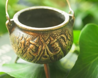 May include: A small, round, metallic bronze-toned cauldron featuring three short legs and a thin, arched handle. The exterior surface is decorated with embossed patterns of pointed hats and crescent moons, creating a textured finish.
