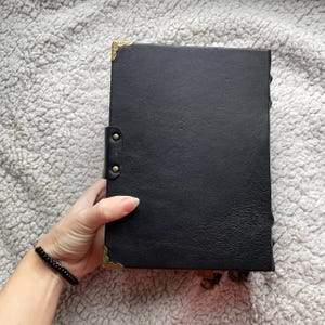 May include: A black leather journal with gold-coloured corner protectors and clasps. The journal is held in a hand, and the cover has a textured appearance. The journal is closed with a leather strap and brass-coloured studs.
