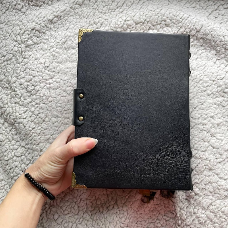 May include: A black leather journal with gold-coloured corner protectors and clasps. The journal is held in a hand, and the cover has a textured appearance. The journal is closed with a leather strap and brass-coloured studs.