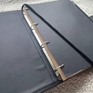 May include: An open black binder with silver metal rings and a black elastic band closure. The pages are a dark gray color, and the spine has a wooden appearance. The binder is lying on a soft, light-colored surface.