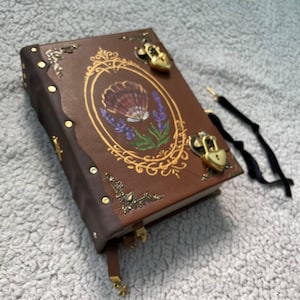 Könnte beinhalten: Ein braunes, in Leder gebundenes Tagebuch mit einem goldfarbenen ovalen Rahmen, der eine Illustration von einer Muschel und Lavendel enthält. Der Einband ist mit verzierten goldfarbenen Metallecken und zwei herzförmigen Vorhängeschlössern versehen.