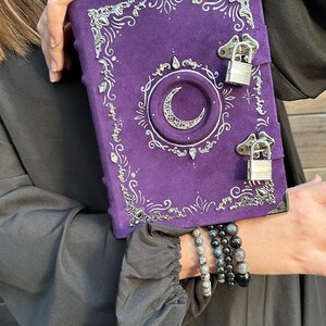May include: A purple journal with silver detailing, featuring a crescent moon and decorative corners. The journal has two silver clasps and a textured cover. The design is gothic and mystical, with a suede-like finish.