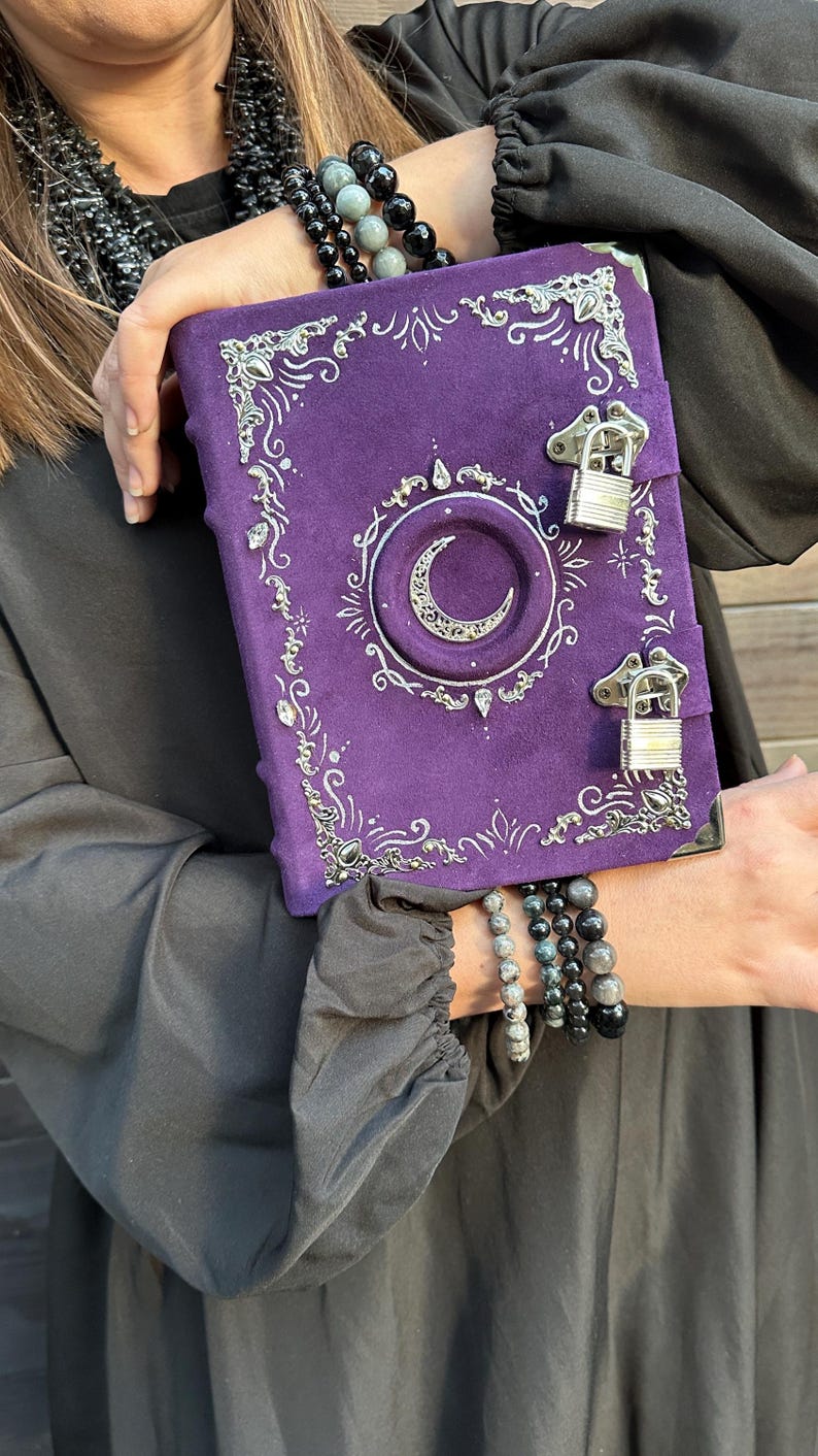 May include: A purple journal with silver detailing, featuring a crescent moon and decorative corners. The journal has two silver clasps and a textured cover. The design is gothic and mystical, with a suede-like finish.