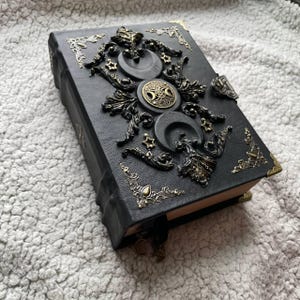 May include: Black leather journal with bronze-toned decorative elements. The cover is adorned with crescent moons, stars, and a central circular emblem. Bronze corner protectors and a ribbon bookmark enhance the design. A gothic-style notebook.