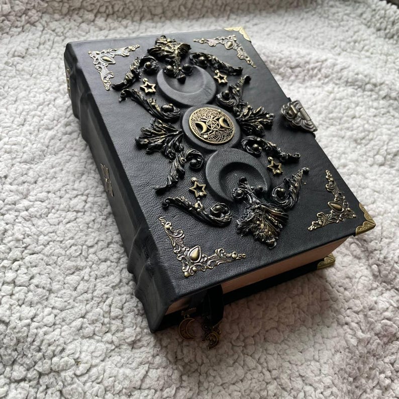 May include: Black leather journal with bronze-toned decorative elements. The cover is adorned with crescent moons, stars, and a central circular emblem. Bronze corner protectors and a ribbon bookmark enhance the design. A gothic-style notebook.