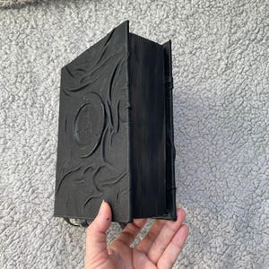 May include: A black, textured book with a circular emblem on the cover. The book has a detailed, embossed design and is held by a hand. The book's spine is visible, and the pages are closed.