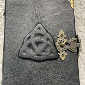May include: Black leather journal with a raised Celtic knot design and antique brass clasp. The journal has a textured surface and decorative corner accents. The clasp is ornate and secures the journal closed.