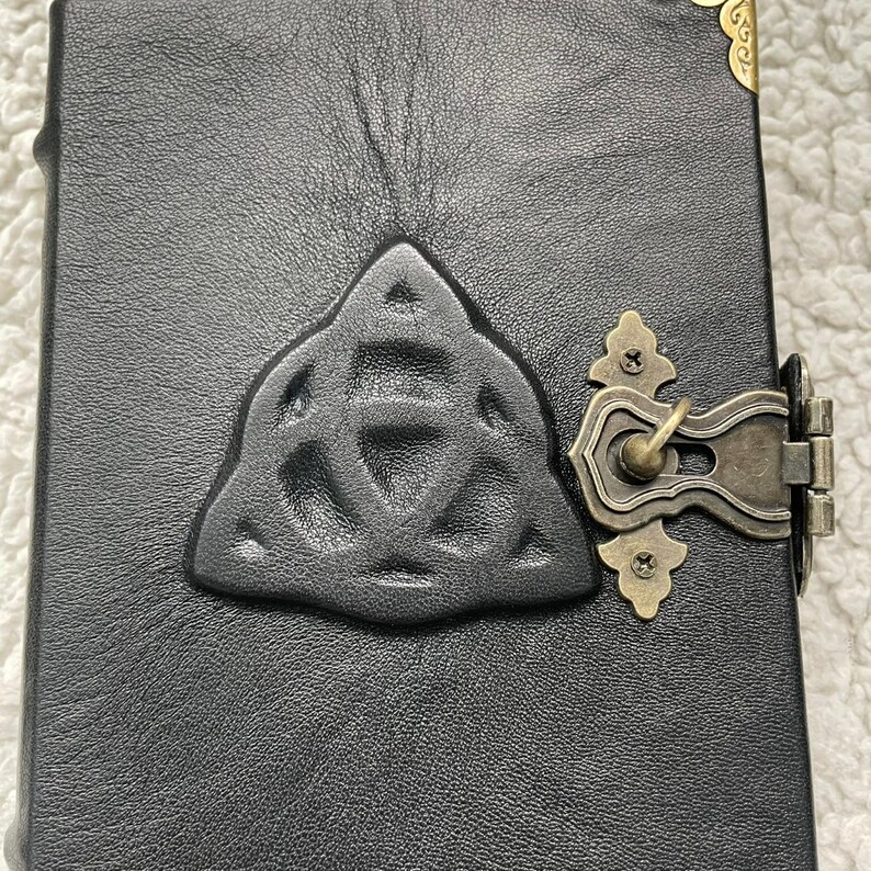 May include: Black leather journal with a raised Celtic knot design and antique brass clasp. The journal has a textured surface and decorative corner accents. The clasp is ornate and secures the journal closed.