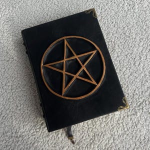 May include: A black leather-bound journal with a gold pentagram design on the front cover. The journal has a brass clasp and a ribbon bookmark.
