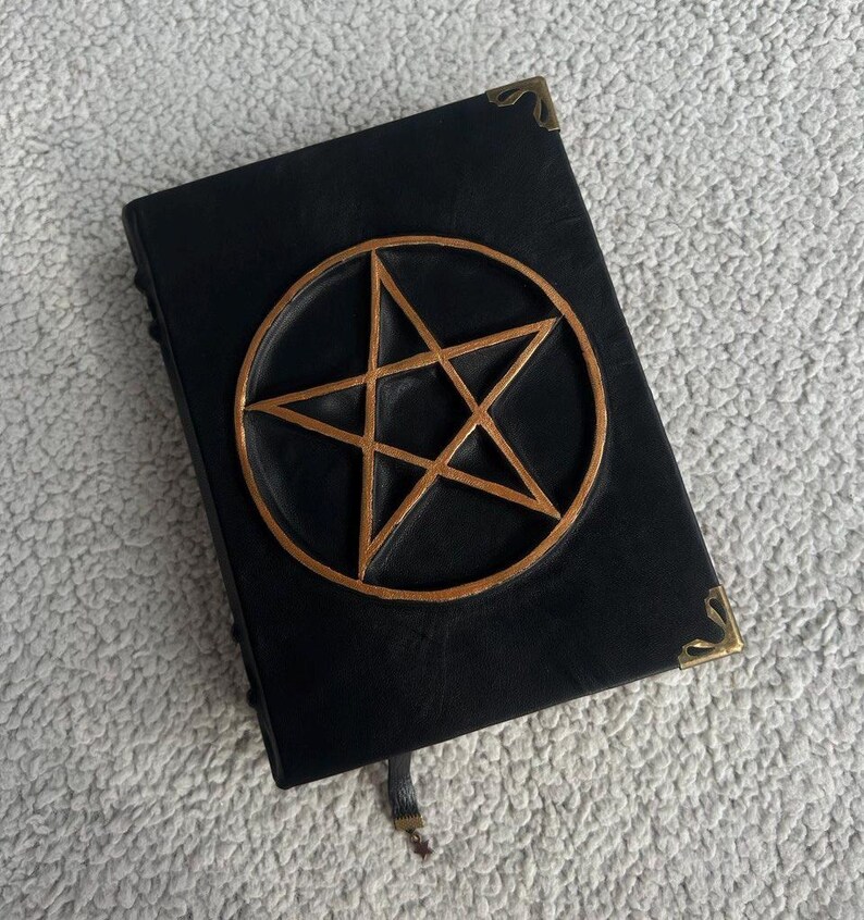 May include: A black leather-bound journal with a gold pentagram design on the front cover. The journal has a brass clasp and a ribbon bookmark.