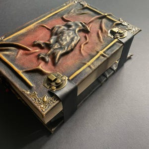 K&ouml;nnte beinhalten: Ein handgefertigtes Lederjournal mit einem erhabenen Rabenmuster auf dem Einband. Das Journal hat einen Verschluss aus schwarzem Leder und dekorative Metallecken. Der Einband ist eine Mischung aus Schwarz, Braun und Rot.