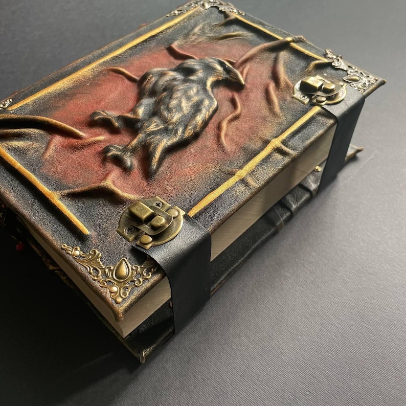 K&ouml;nnte beinhalten: Ein handgefertigtes Lederjournal mit einem erhabenen Rabenmuster auf dem Einband. Das Journal hat einen Verschluss aus schwarzem Leder und dekorative Metallecken. Der Einband ist eine Mischung aus Schwarz, Braun und Rot.