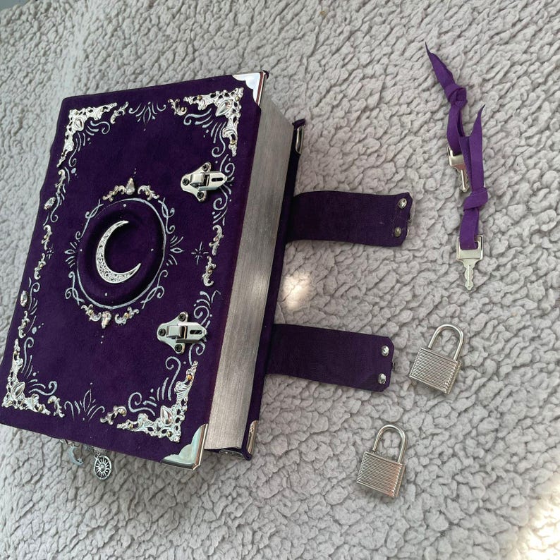 May include: A purple journal with silver detailing, a crescent moon design, and metal clasps. Includes a purple strap with keys and two silver padlocks. The journal's cover has ornate silver detailing.