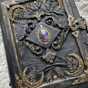 May include: A black leather journal with ornate gold-colored metal accents. The cover features embossed leaf and crescent moon designs, a central purple gemstone, and a metal clasp. The edges of the journal are also trimmed with gold-colored metal.