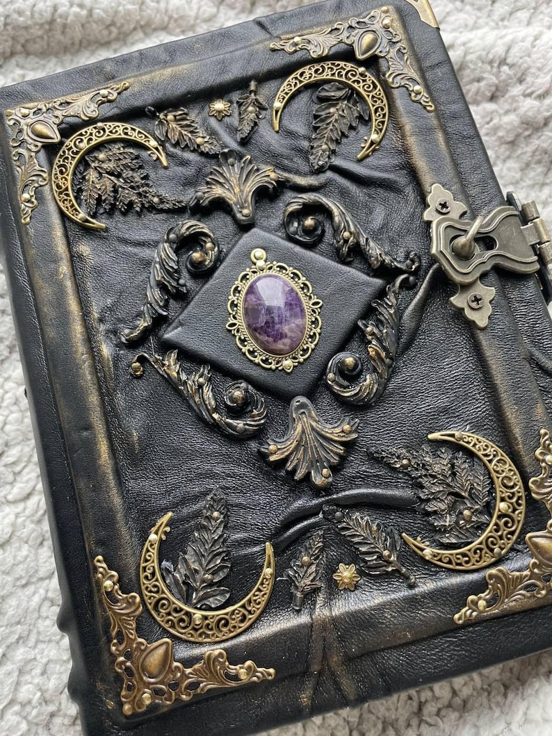 May include: A black leather journal with ornate gold-colored metal accents. The cover features embossed leaf and crescent moon designs, a central purple gemstone, and a metal clasp. The edges of the journal are also trimmed with gold-colored metal.