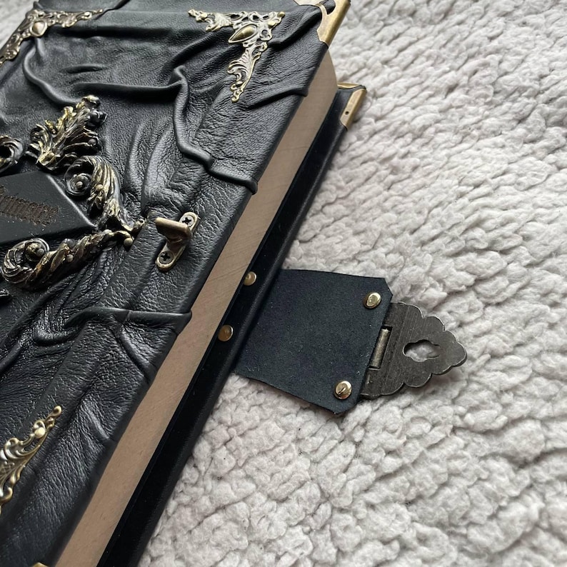May include: A black leather-bound journal with ornate brass corner accents and a clasp closure. The cover features embossed designs and decorative metal embellishments. The journal's pages are visible along the edge, and the overall aesthetic is vintage and gothic.