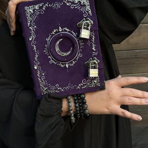 May include: A purple journal with silver detailing and a crescent moon motif. The journal features two small padlocks and decorative silver filigree. The journal is held against a black backdrop.