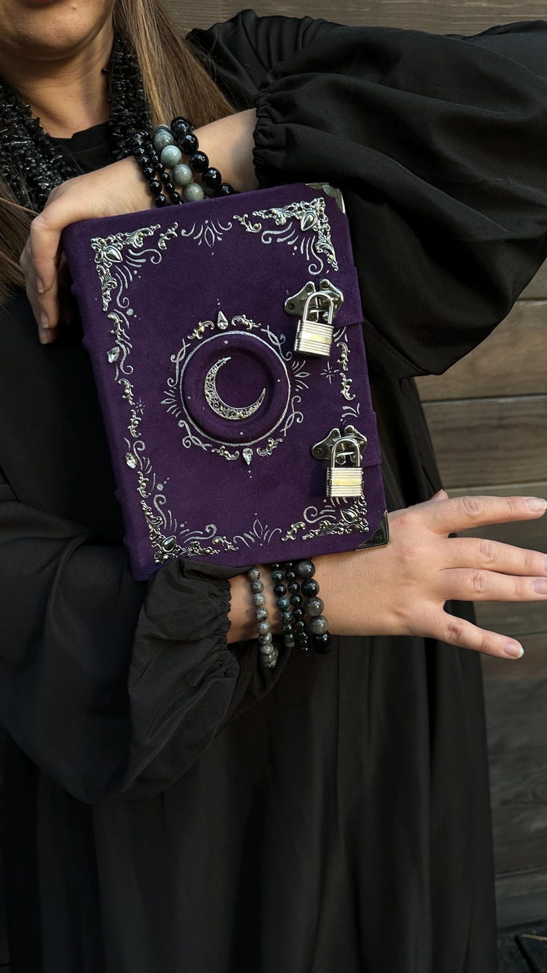 May include: A purple journal with silver detailing and a crescent moon motif. The journal features two small padlocks and decorative silver filigree. The journal is held against a black backdrop.