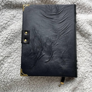 May include: A black leather journal with a textured surface, gold corner protectors, and a snap closure. A black ribbon with a gold star charm serves as a bookmark. The journal is closed and resting on a soft, gray surface.