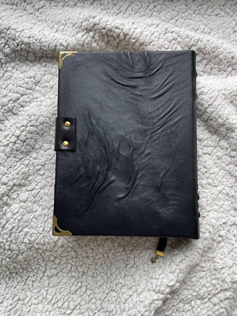 May include: A black leather journal with a textured surface, gold corner protectors, and a snap closure. A black ribbon with a gold star charm serves as a bookmark. The journal is closed and resting on a soft, gray surface.