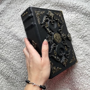 May include: A black, ornate book with gold-coloured embellishments. The cover features intricate relief designs, including circular and floral motifs. The book's edges are also adorned with gold accents. The book is held in a hand.