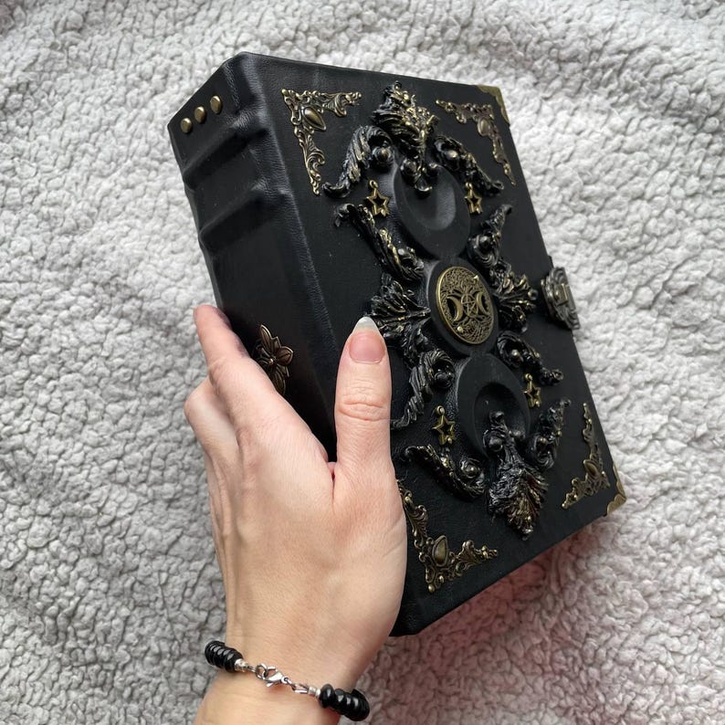 May include: A black, ornate book with gold-coloured embellishments. The cover features intricate relief designs, including circular and floral motifs. The book's edges are also adorned with gold accents. The book is held in a hand.